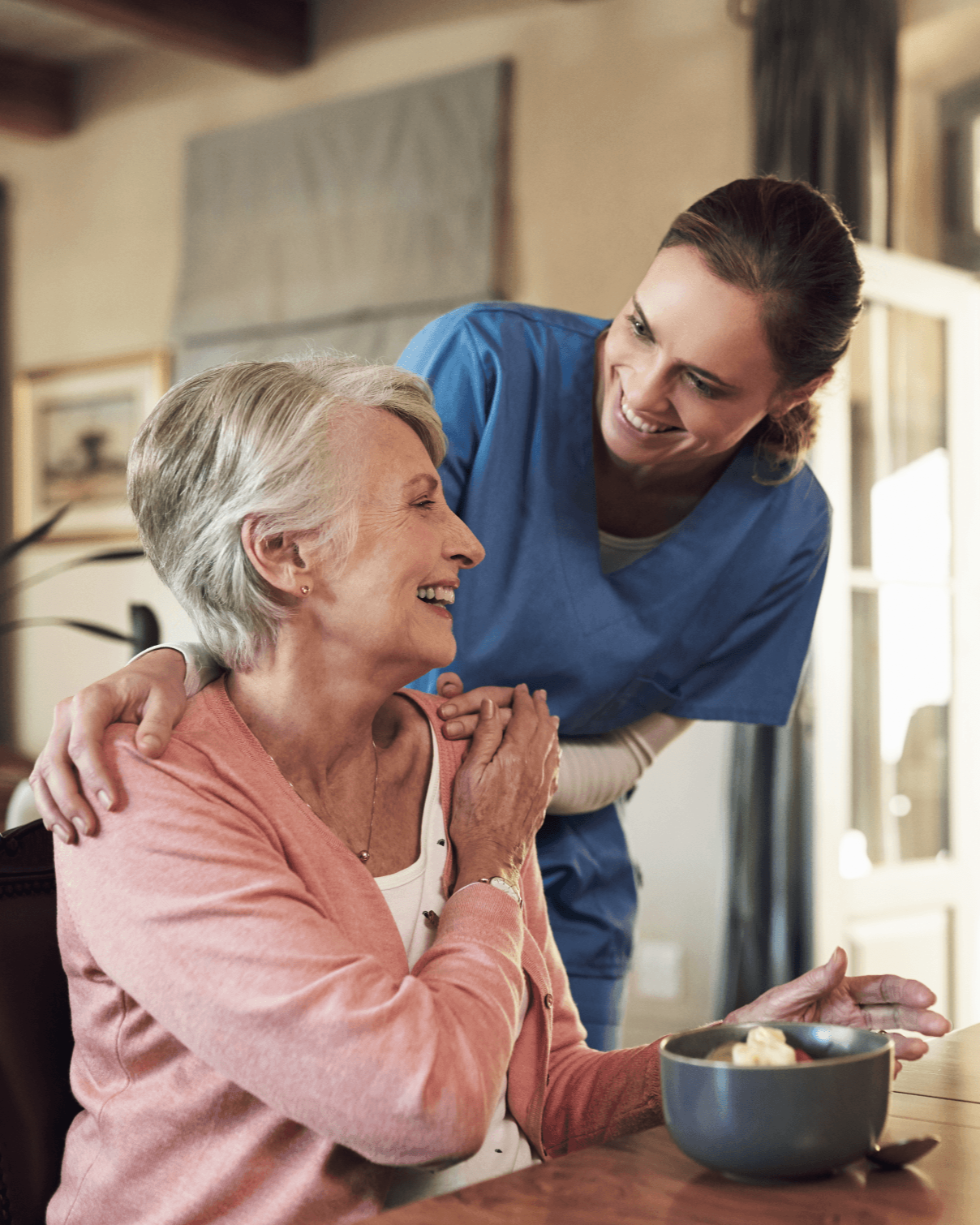 A friendly caregiver in blue scrubs gently supports and smiles with an elderly woman seated at a dining table in a bright home setting.