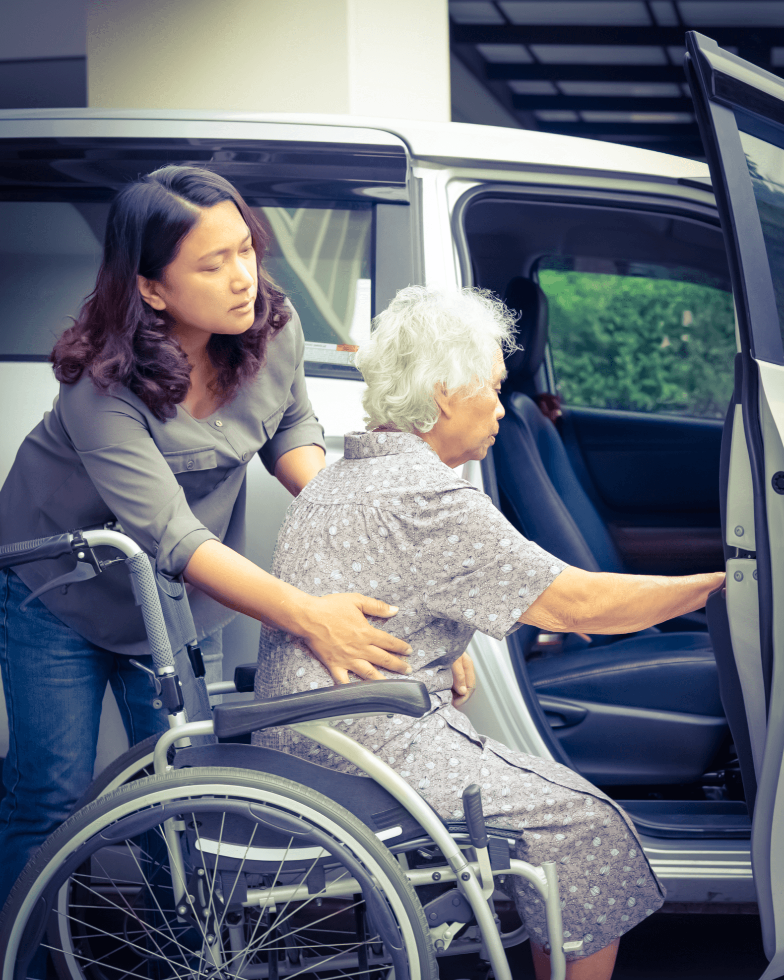 Caregiver helping an elderly woman in a wheelchair transfer safely into a white vehicle.