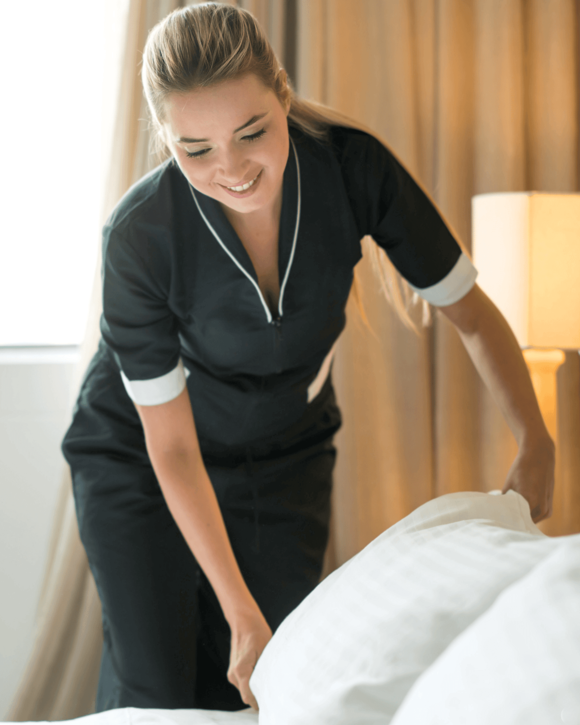 Caregiver smiling while making a bed in a warmly lit room.
